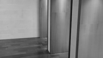 An interior space with wooden panels and a wooden floor. The lighting creates soft shadows, emphasizing the texture of the materials. The composition is minimalist and geometric, focusing on lines and surfaces.