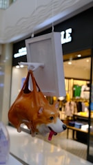 A hanging ceramic sculpture resembling a corgi dog, designed to look like it's being held by a handle attached to a square panel. The background shows a blurred view of a clothing store interior.