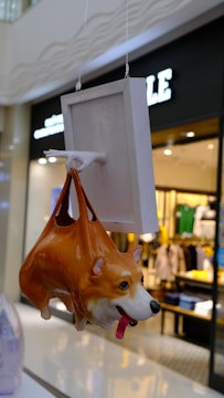 A hanging ceramic sculpture resembling a corgi dog, designed to look like it's being held by a handle attached to a square panel. The background shows a blurred view of a clothing store interior.