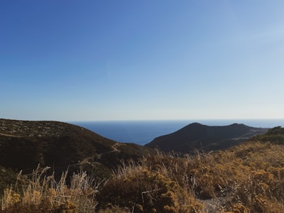 Rolling hills covered with dry grass lead to a view of the ocean horizon under a clear blue sky.