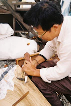 A person is engaged in woodworking, holding a carving tool to shape the surface of a wooden piece. The setting appears to be outdoors, with a tree in the background and a blanket or cloth nearby. The person is focused on their craft.
