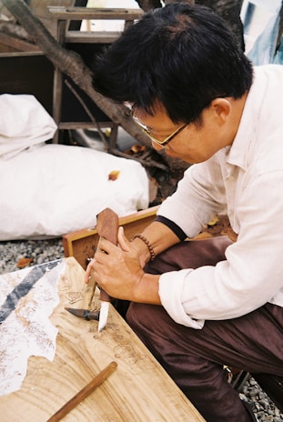 A person is engaged in woodworking, holding a carving tool to shape the surface of a wooden piece. The setting appears to be outdoors, with a tree in the background and a blanket or cloth nearby. The person is focused on their craft.