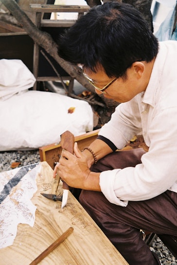 A person is engaged in woodworking, holding a carving tool to shape the surface of a wooden piece. The setting appears to be outdoors, with a tree in the background and a blanket or cloth nearby. The person is focused on their craft.
