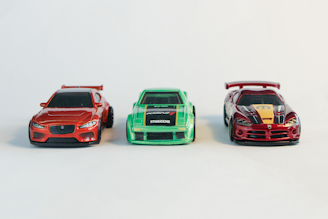 A trio of detailed toy cars in vibrant colors is placed against a plain, light background. On the left, there is a red sports car with sleek lines and sharp features. In the middle, a green toy car displays bold racing stripes and logos. On the right, a red and black car with a prominent rear wing and distinctive design elements attracts attention.