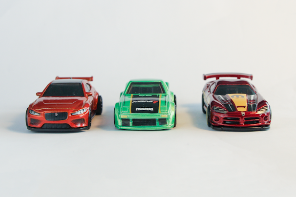 A trio of detailed toy cars in vibrant colors is placed against a plain, light background. On the left, there is a red sports car with sleek lines and sharp features. In the middle, a green toy car displays bold racing stripes and logos. On the right, a red and black car with a prominent rear wing and distinctive design elements attracts attention.