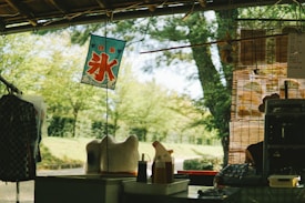 An outdoor scene resembling a quaint stall with traditional Japanese elements. There is a decorative hanging sign with red kanji characters, and various items such as a patterned cloth, bottles, and a small decorative object resembling a cat. The background features green trees and bushes, suggesting a park or garden setting.