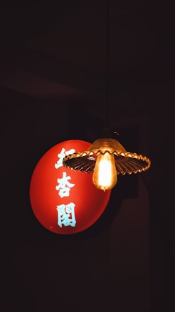 A vintage lightbulb with a decorative fixture hangs in front of a red circular sign with white Chinese characters on it, set against a dark background.
