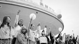 A group of people stand together, raising their fists in a gesture of solidarity and protest. One person is holding a flower, while another holds a balloon. They appear to be gathered in front of a large, curved building.