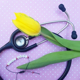A yellow tulip lays across a stethoscope on a pink background. The background is patterned with small white hearts. Below the stethoscope, there is a bracelet with beads spelling out 'stay safe'.