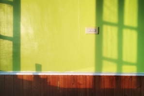 A brightly lit green wall features a three-button light switch. Shadows from a window pattern stretch across the surface, creating geometric shapes. At the bottom, a brown wooden panel forms the lower section of the wall.