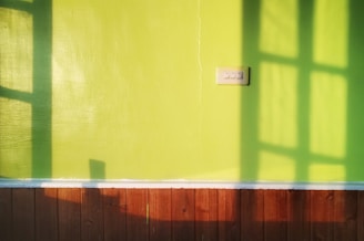 A brightly lit green wall features a three-button light switch. Shadows from a window pattern stretch across the surface, creating geometric shapes. At the bottom, a brown wooden panel forms the lower section of the wall.