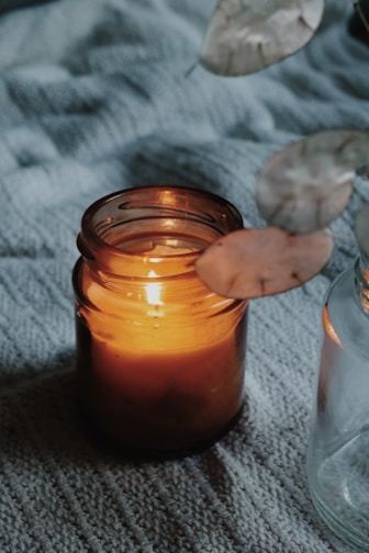 Close-up of a frosted glass jar candle glowing softly on a wooden table with dried flowers nearby.
