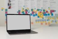 A laptop with a blank screen sits on a white desk. In the background, multiple colorful sticky notes and documents are posted on a large board, suggesting a creative or collaborative workspace environment.