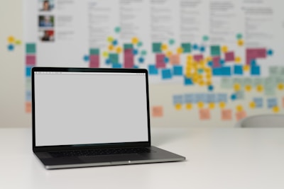 A laptop with a blank screen sits on a white desk. In the background, multiple colorful sticky notes and documents are posted on a large board, suggesting a creative or collaborative workspace environment.