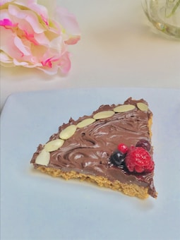 A slice of dessert with a chocolate topping and garnished with almonds and fresh berries, laid on a white plate. Soft pink and white flowers are placed to the side, creating a delicate and elegant presentation.
