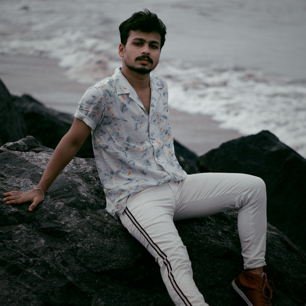 man in white and red floral button up shirt and white pants sitting on rock