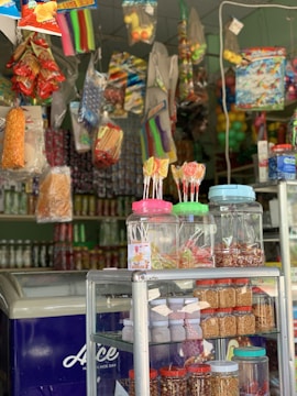 A vibrant store interior features an assortment of colorful packaged snacks and candy hanging from the ceiling. There are jars of lollipops and various nuts displayed on a glass shelf. The freezer at the bottom left corner has the word 'Ice' in white letters on a blue background.