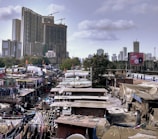 A bustling urban landscape with a large contrast between a high-rise building under construction and a densely populated informal settlement. Clothes hang drying on lines between makeshift structures, and a train passes by in the background. A large billboard stands out amidst the varied structures, framed by sparse greenery and a cloudy sky.