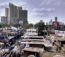 A bustling urban landscape with a large contrast between a high-rise building under construction and a densely populated informal settlement. Clothes hang drying on lines between makeshift structures, and a train passes by in the background. A large billboard stands out amidst the varied structures, framed by sparse greenery and a cloudy sky.