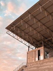 A modern, industrial-style building with a large overhanging metal roof structure. The structure is supported by a grid of steel beams and columns. Below the roof, there is a building with large glass windows reflecting a soft peach and blue sky. The setting appears to be an open, outdoor area.