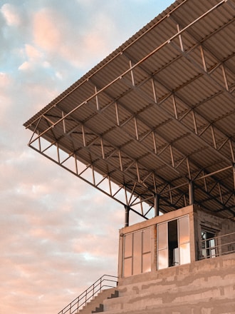 A modern, industrial-style building with a large overhanging metal roof structure. The structure is supported by a grid of steel beams and columns. Below the roof, there is a building with large glass windows reflecting a soft peach and blue sky. The setting appears to be an open, outdoor area.