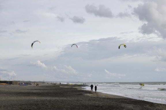 A cloudy beach scene with several kitesurfers in the distance and three kites visible in the sky. People are walking along the sandy shore as waves gently crash onto the beach.