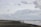 A cloudy beach scene with several kitesurfers in the distance and three kites visible in the sky. People are walking along the sandy shore as waves gently crash onto the beach.