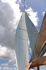 Tall sails stretch upward against a backdrop of partly cloudy sky. The sails are crisp and white, contrasting sharply with the blue sky. The angle of the photograph creates a dynamic perspective, emphasizing the height and elegance of the sailboat's masts. The edge of the boat is visible in the lower corner, with a muted brown color.