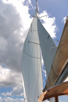 Tall sails stretch upward against a backdrop of partly cloudy sky. The sails are crisp and white, contrasting sharply with the blue sky. The angle of the photograph creates a dynamic perspective, emphasizing the height and elegance of the sailboat's masts. The edge of the boat is visible in the lower corner, with a muted brown color.