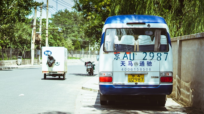 A small white and blue van is parked on the side of a tree-lined street. The van has a license plate and phone number written in blue on the rear. Nearby, a small delivery truck with Chinese characters is driving down the road, and a motorcycle is parked on the curb. The street is flanked by a tall wall and utility poles with overhead wires.