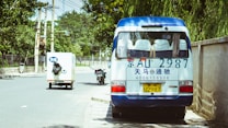 A small white and blue van is parked on the side of a tree-lined street. The van has a license plate and phone number written in blue on the rear. Nearby, a small delivery truck with Chinese characters is driving down the road, and a motorcycle is parked on the curb. The street is flanked by a tall wall and utility poles with overhead wires.
