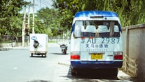 A small white and blue van is parked on the side of a tree-lined street. The van has a license plate and phone number written in blue on the rear. Nearby, a small delivery truck with Chinese characters is driving down the road, and a motorcycle is parked on the curb. The street is flanked by a tall wall and utility poles with overhead wires.