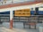 A railway station sign in both Hindi and English indicates 'Ujjain Junction.' The yellow sign is prominently placed on a white and red painted column. A metal bench is situated in front of the sign along with a section of railway tracks and a blue train visible in the background. Part of a footbridge and a person's legs in motion can be seen above.