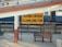 A railway station sign in both Hindi and English indicates 'Ujjain Junction.' The yellow sign is prominently placed on a white and red painted column. A metal bench is situated in front of the sign along with a section of railway tracks and a blue train visible in the background. Part of a footbridge and a person's legs in motion can be seen above.