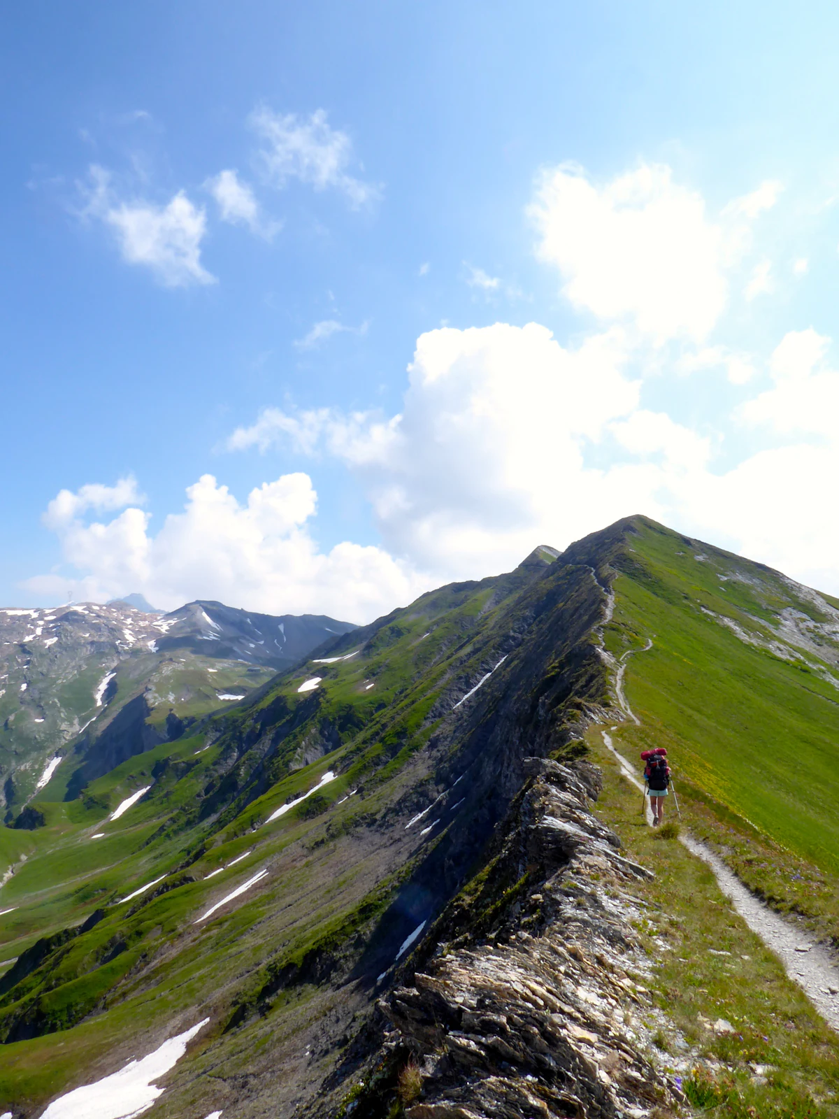 A hiker on a narrow grassy ridge path in the Beaufortain, with rocky peaks behind