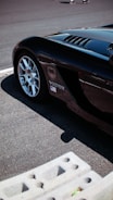 A portion of a sleek black sports car is parked on a track with part of its front wheel visible. There is a Viper SRT-10 logo on the side, as well as an American flag sticker. The background shows a textured pavement and the image is taken in bright sunlight, casting shadows.