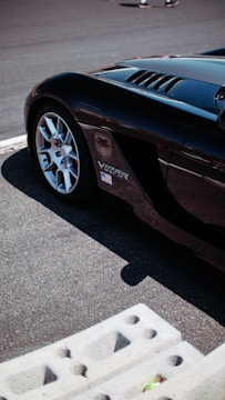 A portion of a sleek black sports car is parked on a track with part of its front wheel visible. There is a Viper SRT-10 logo on the side, as well as an American flag sticker. The background shows a textured pavement and the image is taken in bright sunlight, casting shadows.