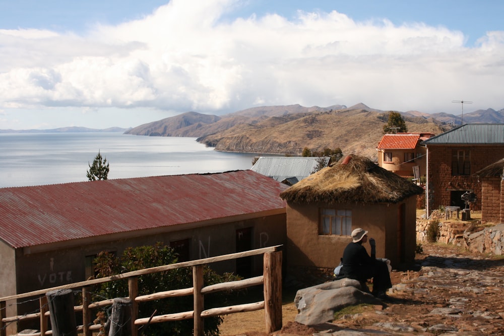 man in black jacket sitting on brown wooden fence near body of water during daytime