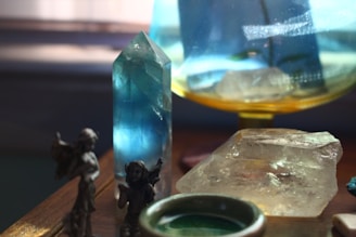 A collection of crystals and gemstones displayed on a wooden surface, including a prominent tall blue crystal and a large clear quartz. Small figurines are also visible, adding a mystical and decorative element. Soft lighting enhances the colors and transparency of the stones.