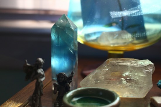A collection of crystals and gemstones displayed on a wooden surface, including a prominent tall blue crystal and a large clear quartz. Small figurines are also visible, adding a mystical and decorative element. Soft lighting enhances the colors and transparency of the stones.