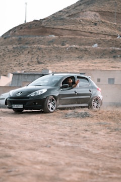 A person is sitting in a black Peugeot car parked on a rough, sandy terrain. The background features a hill with sparse vegetation and a few buildings at the foothill.