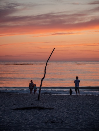 A serene beach scene during sunset with a warm, colorful sky transitioning from orange to pink hues. Two adults and a child stand at the edge of the gently lapping waves, silhouetted against the vivid backdrop. A lone stick protrudes from the sand, adding an element of natural simplicity. The overall scene conveys a sense of peace and introspection.