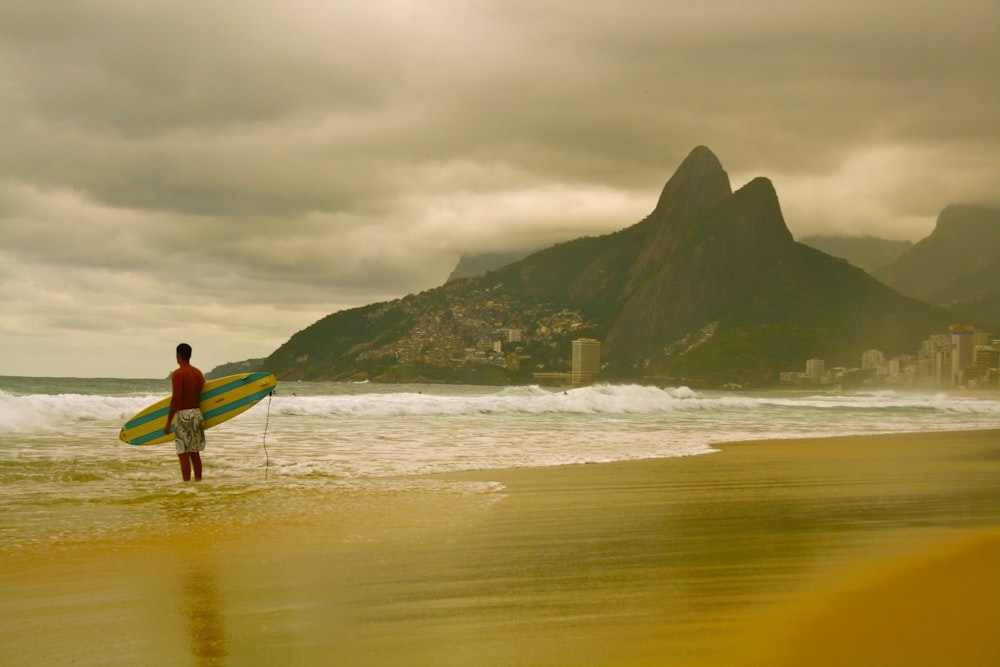 빨간 셔츠와 갈색 반바지를 입은 남자가 낮에 해변을 걷는 흰색 서핑보드를 들고 있다