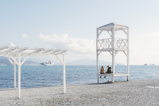 woman in blue shirt sitting on white wooden bench near body of water during daytime in Novorossiysk Russia