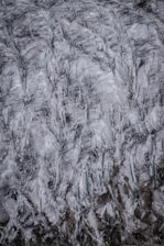 An aerial view of a glacier showing intricate patterns and textures in shades of white, gray, and black. The surface of the glacier appears to be heavily crevassed, with deep cracks and rugged lines.