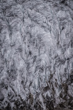 An aerial view of a glacier showing intricate patterns and textures in shades of white, gray, and black. The surface of the glacier appears to be heavily crevassed, with deep cracks and rugged lines.
