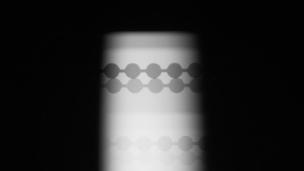 A vertical pattern of circular shapes aligned in rows and columns is cast as a shadow. The pattern is surrounded by a dark background, highlighting the contrast between light and shadow.