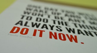 A motivational quote is printed on a piece of paper. The text reads 'One day you will wake up and there won’t be any more time to do the things you always wanted. Do it now.' The words 'Do it now' are emphasized in bold red letters. The rest of the text is in black, and the quote is attributed to Paulo Coelho.