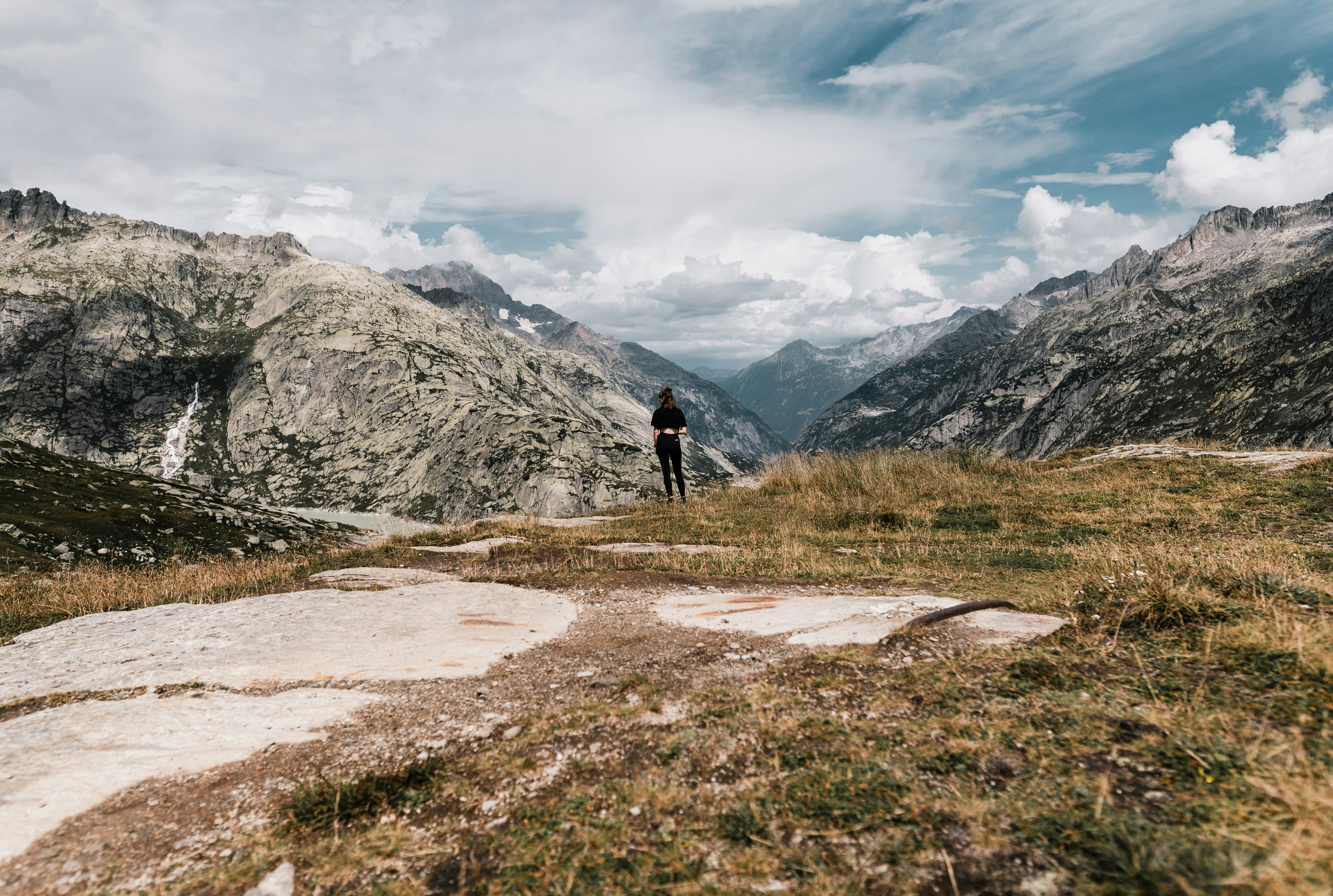 person in black jacket walking on dirt road near rocky mountain under white cloudy sky during