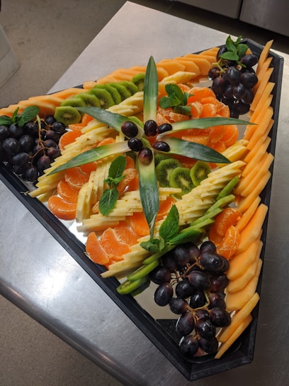 A colorful, elegant fruit platter featuring sliced melons, berries, and grapes arranged neatly on a wooden board.